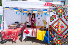 Two people stand behind a table decorated with Ukrainian flags. The table is covered with traditional fabrics and tableware. A panel with a colorful pattern stands nearby.