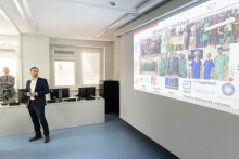 A presenter stands by the screen and holds a presentation remote. The slide shows the title “Scientific Activity” and photos of a team.