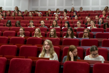 Large audience seated in rows of red chairs in a lecture hall. All attendees are facing forward toward the stage