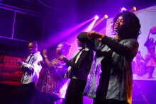 Four performers on stage moving rhythmically in a line, illuminated by purple and pink stage lights.