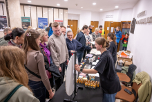 A crowded educational stand in a museum space. The presenter demonstrates materials at a table covered with numerous small bottles, leaflets, and tools, while a group of visitors gathers in front of the stand.