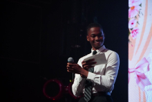 A person standing on stage holding a microphone and reading from a sheet of paper during an event.
