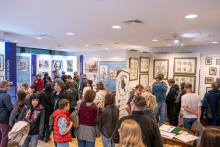 A museum exhibition hall filled with visitors. Numerous people view anatomical charts and exhibits arranged on walls and at educational stations.