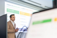 A presenter stands next to the screen and holds a presentation remote. The slide shows the title “Where AI Works Today – and Where It Will in the Future.”