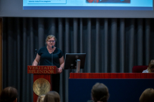 Person standing at a podium with microphones and a computer monitor, delivering a talk in front of a dark curtain backdrop.