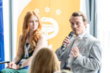 One person is seated holding a microphone; another person is seated nearby, with a yellow banner in the background.