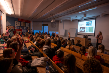 A lecture in an auditorium filled with attendees. People sit on benches and raise their hands, while at the front a presenter displays an educational slide on a screen.