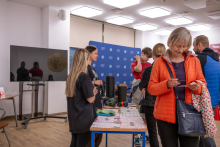 An informational and educational stand in a bright room. Visitors stand at a table with leaflets and teaching aids, talking with presenters; a backdrop with logos and a screen is visible behind them.