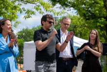 Four people stand side by side. One person holds a microphone and speaks, while the others stand nearby and listen. Greenery and a tent are visible in the background.