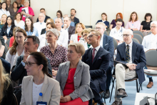The audience sits in rows of chairs and listens to the presentation. Some people hold notebooks or documents.