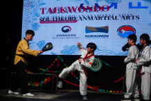 Children performing a taekwondo demonstration on a stage, with one child executing a kick toward a training pad held by an instructor. A large screen in the background displays the text “Taekwondo – South Korean Martial Arts” and official logos.