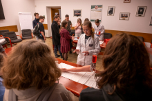 Educational workshops in a museum room. At tables, several people observe a demonstration using an arm model with a transparent container filled with red liquid, while the presenter explains the course of the experiment.