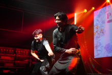 Two performers on a stage execute a synchronized martial arts sequence, with extended arm movements, under red stage lighting and in front of a decorative backdrop.