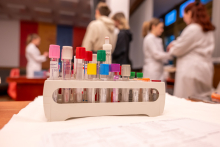A rack with laboratory test tubes with colored caps placed on a table. In the blurred background, people in lab coats are visible during educational activities.