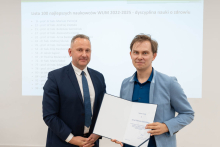 Two men are posing for a photo. One of them is holding an open diploma in his hands.
