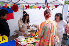 Four people stand at a table with food. One person is serving a dish onto a plate. Dumplings and other dishes are on the table. Colorful garlands and a flag hang in the background.