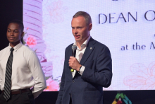 Two adults standing on a stage during a formal event; one person speaks into a handheld microphone while a presentation slide with text is displayed on a large screen behind them.