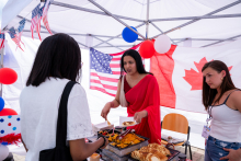Three people stand at a food station. One person serves a portion from a warmer. Crepes and toppings are on the table. In the background, the flags of the United States and Canada are visible.