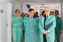 A group of people walks along a hospital corridor wearing green disposable protective gowns. One person is holding a document.