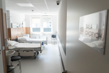 A hospital room with two beds, each equipped with medical equipment. The room is bright, with a window and a chair placed by the wall.