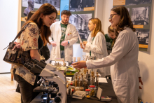 A workshop station with a microscope and sample containers. Presenters in lab coats show laboratory materials to visitors, who observe the exhibits and talk at the table.