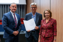 Two men and a woman are posing together for a photo. The man in the middle is holding an open diploma in his hands.