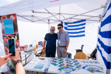 Two people stand by a table with leaflets and promotional materials. A third person is taking a photo with a phone. In the background, the Greek flag and a poster reading “Greece” are visible.
