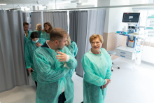 A group of people in green protective gowns stands in a medical ward and talks. In the background, curtains separating stations and medical equipment are visible.