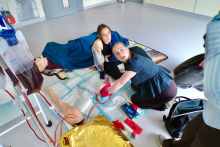 Dwie osoby udzielają pomocy medycznej podczas symulacji na podłodze sali dydaktycznej. Jedna osoba trzyma w rękach gaziki i wykonuje czynności przy fantomie kończyny dolnej, a druga osoba opiera się obok. Obok leżą elementy sprzętu medycznego, w tym torby z płynem, zestawy opatrunkowe oraz złota folia termoizolacyjna.