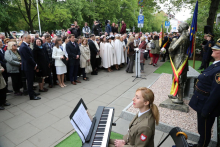 Uroczystość plenerowa przy pomniku Stanisławy Leszczyńskiej. Przed pomnikiem stoi mównica z mikrofonem. Liczna grupa uczestników zgromadzona jest wokół, część osób trzyma dokumenty lub torebki. Na pierwszym planie znajduje się instrument klawiszowy z nutami na pulpicie.