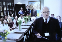 A formal meeting room with multiple people seated around a long table. One person standing in the foreground is holding a sheet of paper. Flower arrangements in glass vases are placed along the table, and bookshelves are visible behind the seated participants.