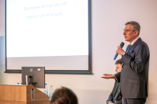 An educational presentation in a lecture hall. A person standing at a lectern speaks into a microphone, while a presentation about the role of laboratories in diagnostics is displayed on the screen behind them.