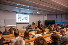 A view of a lecture hall filled with attendees seated on benches. At the front, the presenter discusses a slide, while participants watch the lecture.