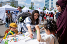 One adult and two children sit at a table and draw. Crayons and sheets of paper are spread out on the table. In the background, tents and people walking around the event can be seen.
