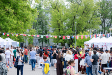 A large group of several dozen people stands in a square between stalls. Flags from various countries hang above them. Trees and buildings are in the background.