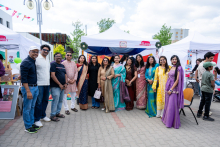 About 14 people stand in a row in front of a tent, posing for a group photo. A stall and decorations are visible in the background.