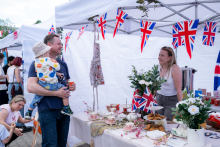 Two people talk at a stall with decorations and tableware. One person is holding a child. Cups, a pitcher, and plates with snacks are on the table.