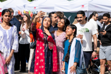 Five people stand close together, taking a photo with a phone. One person is holding the phone with an outstretched arm. A crowd and tents are visible in the background.