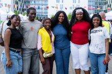 Six people stand side by side and pose for a photo. They stand close together with their arms around each other. Buildings, tents, and strings of flags from various countries are visible in the background.