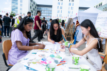 Three people sit at a table and draw on sheets of paper. Crayons, brushes, and cups of water are on the table. In the background, a crowd and stalls set up in the square can be seen.