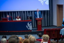 A speaker stands at a lectern labeled “Veritatis Splendor” and addresses the audience. Two people are seated at a table on the stage behind the lectern, with laptops and water bottles placed in front of them.