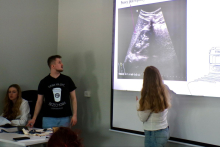 Dwie osoby prowadzą prezentację przy ekranie projekcyjnym z obrazem USG i podpisem „Prawy płat wątroby”. Jedna osoba stoi i wskazuje slajd ręką, a druga osoba siedzi przy stole, na którym znajdują się materiały szkoleniowe oraz drobny sprzęt.