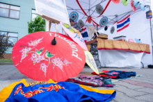 A colorful umbrella with the word “Thailand” lies on the pavement next to laid-out costume elements and fabrics. In the background, there is a stall decorated with ornaments and a flag. One person stands by the table preparing materials.