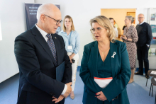 Two people stand facing each other and converse in a bright room. The person on the right is holding a document or a folder with a red edge. The person on the left has their hands folded in front of them. In the background, other people are present, along with a stand holding informational materials. A banner on the wall reads “Medical University of Warsaw.”