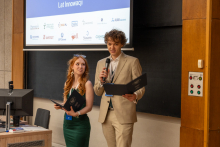 Two people are standing at a lectern in a lecture hall; one person is holding a microphone and a document, the other is holding a folder, with a screen showing partner logos behind them.