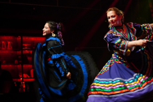 Two performers on stage wearing brightly colored folkloric dresses, captured mid-movement under theatrical lighting.