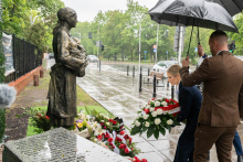 A person places a floral wreath at a memorial statue during rainfall. Other people with umbrellas stand nearby on a wet sidewalk.