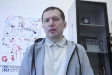 A person standing indoors in front of a wall covered with handwritten notes, drawings, and hearts. A glass cabinet with books is positioned to the right. The person is wearing a light-colored shirt and a hoodie and is facing the camera.