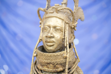 A golden sculpture depicts a human head with an ornate headdress and jewelry elements. The sculpture stands on a pedestal. The background is plain and blurred.