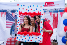 Three people stand close together, holding a large photo frame. They pose for a picture. In the background, the flags of the United States and Canada and stall decorations are visible.